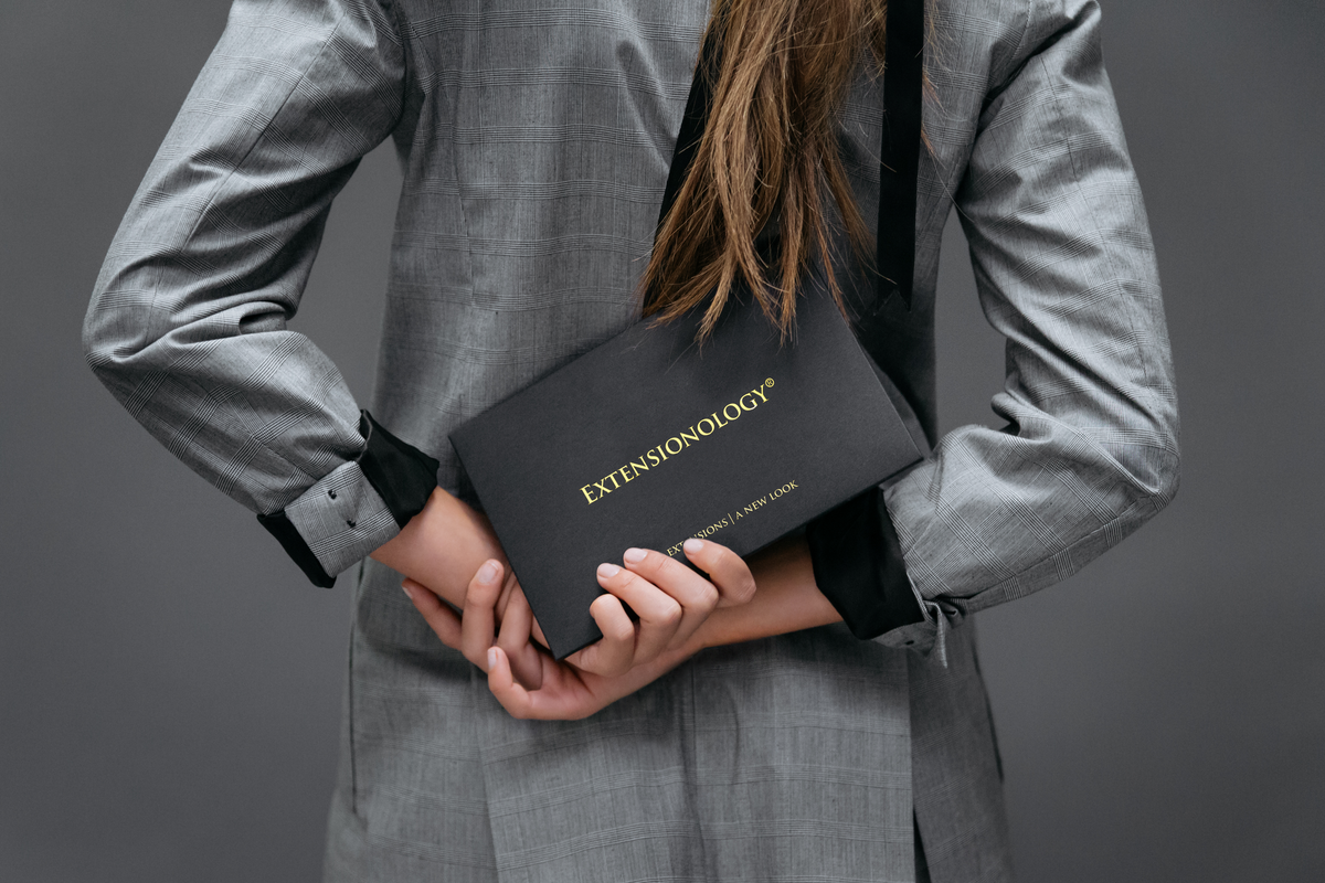 A woman with long brunette hair holding a black Extensionology box behind her back